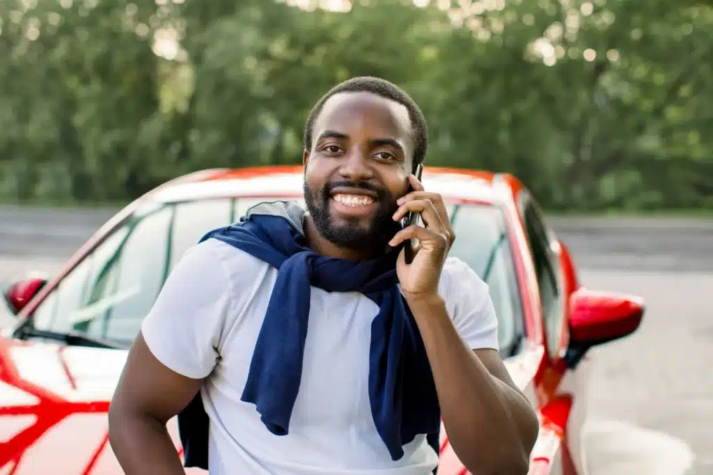 A person smiles and leans on their car while calling a traffic ticket expert for help after receiving a ticket.