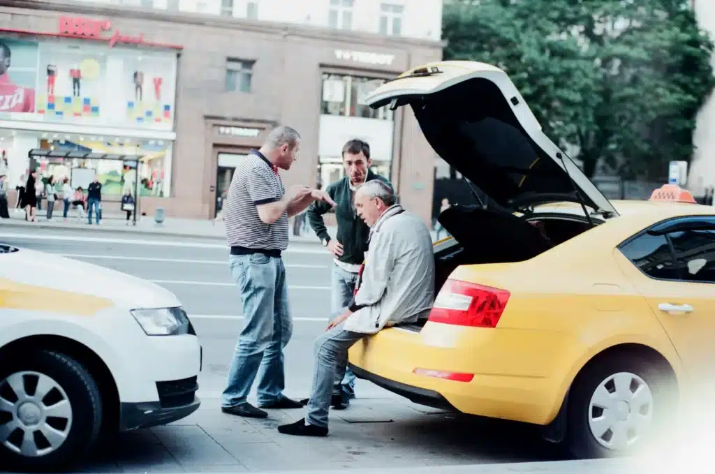 A taxi driver sits in the trunk of their car with the door open after hitting a parked car while two other people talk to them about the accident.